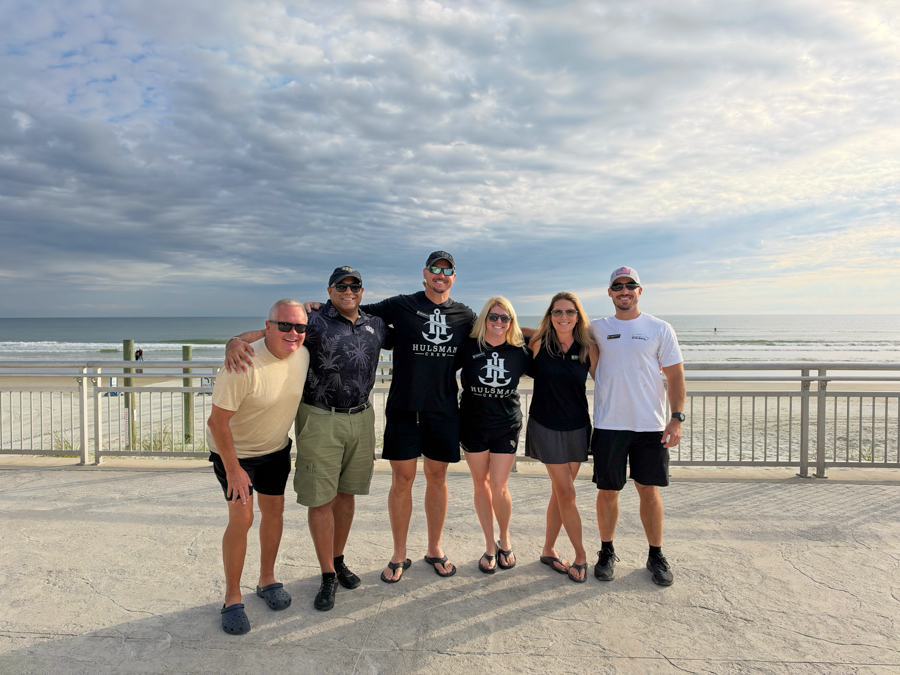 Six adults posing with arms around each other on a beachside boardwalk, ocean and cloudy sky in the background.