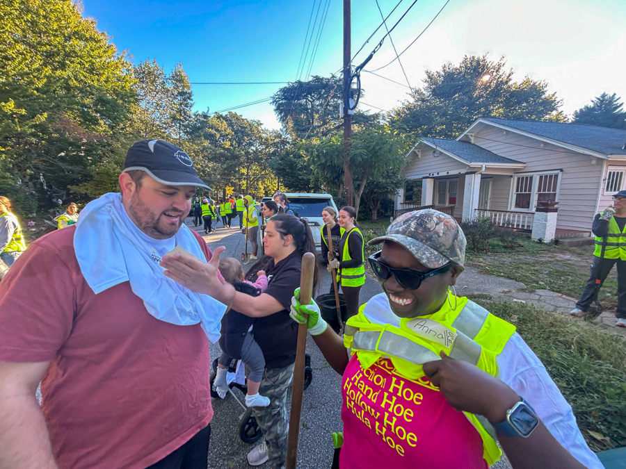 Volunteer group cleaning a suburban street; a man with a towel around his neck chats with a smiling woman in a neon vest.