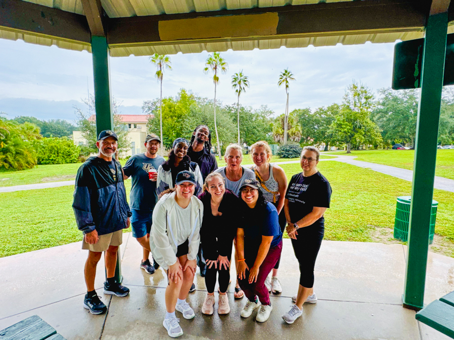 Group of ten people posing together under a park pavilion on a sunny day, with palm trees in the background.