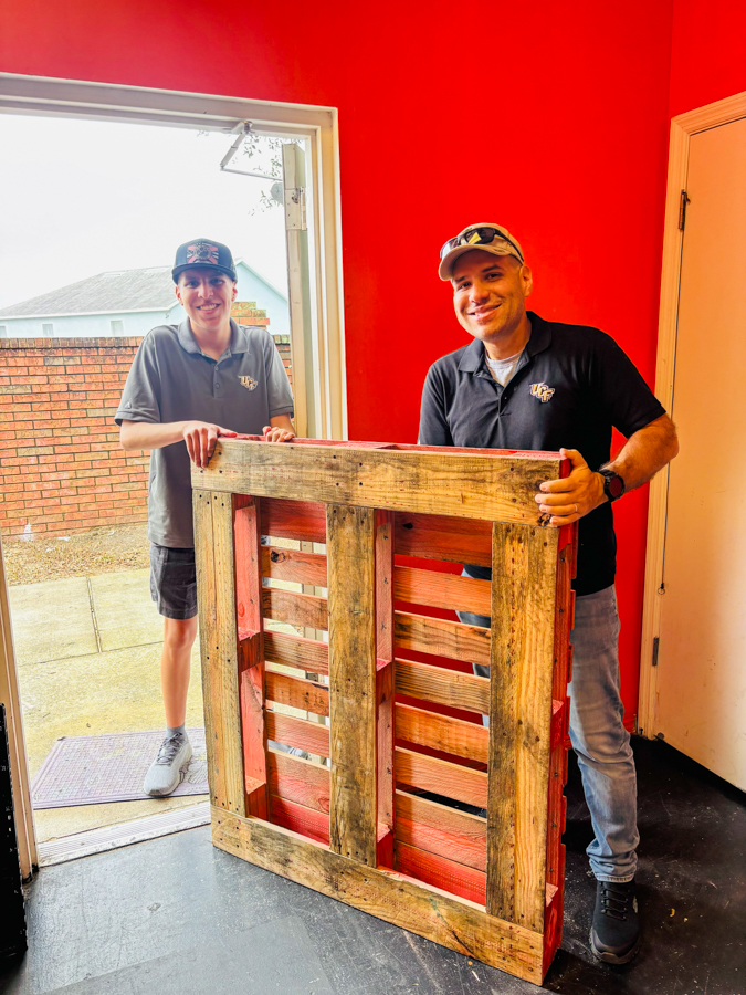 Two men stand in a red-walled room, holding a wooden pallet between them, smiling at the camera inside a building.Outside doorway shows a brick wall and yard.