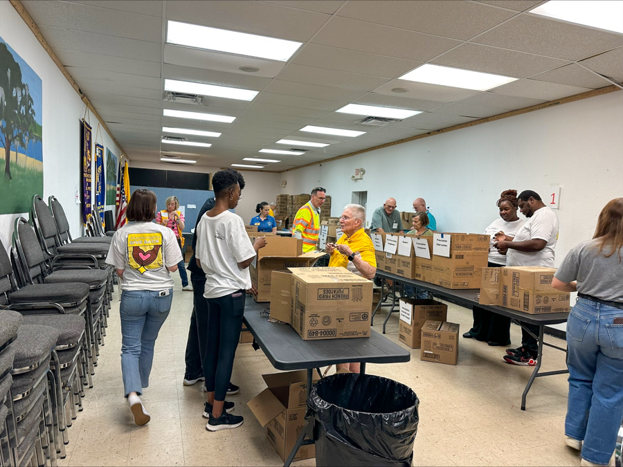 Volunteers in a community hall sorting and packing donated boxes for a charity drive, with tables, boxes, and people working together.