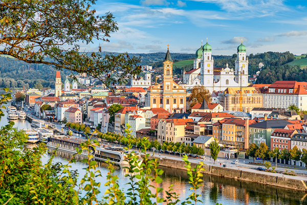 European riverside town with pastel buildings, a large church with green domes, and a tree-framed foreground over a calm river.