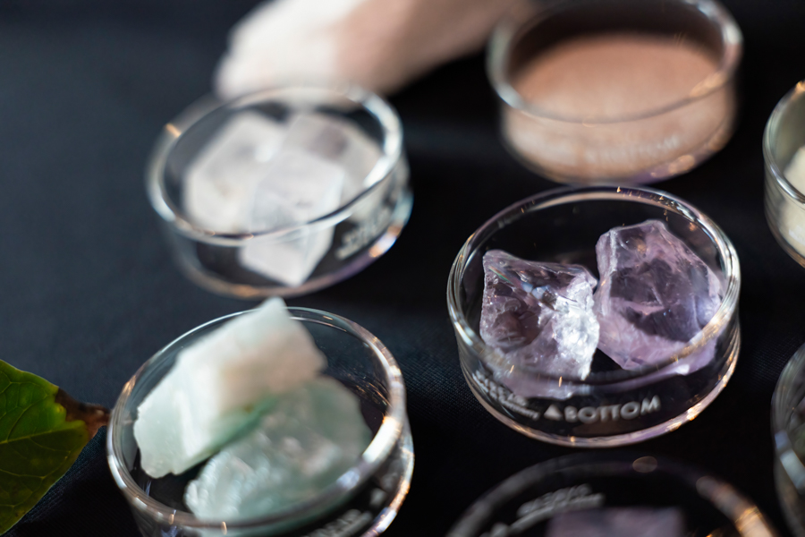Glass bowls filled with assorted crystal chunks on a dark surface; purple amethyst and pale green stones are visible in the foreground and background.