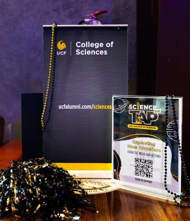 UCF College of Sciences banner on a table with black and gold beads and a smaller Science Tap sign beside it