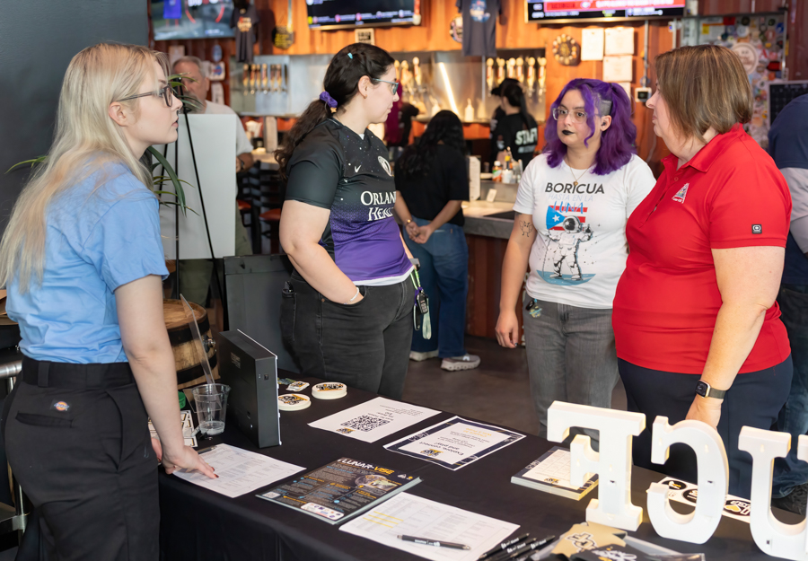 Four women stand around a black booth table with papers and promotional items at a bar or event venue.
