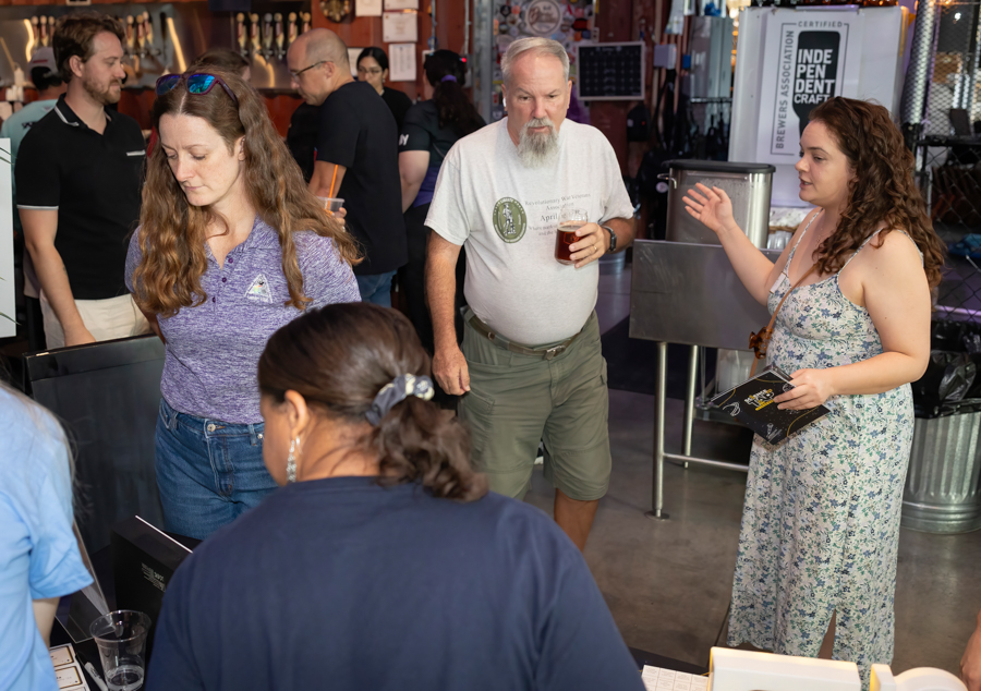 Group of people at a brewery tasting bar; a bearded man holds a beer while a woman in a floral dress talks to staff and others look on.