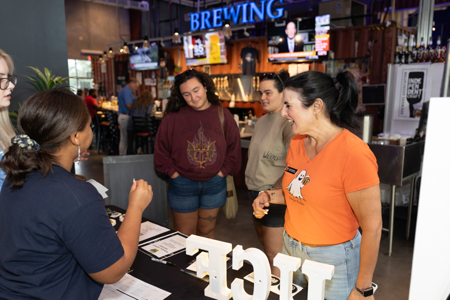 Staff member in an orange shirt chats with a small group at a check-in table inside a brewery, with a 'BREWING' sign overhead.