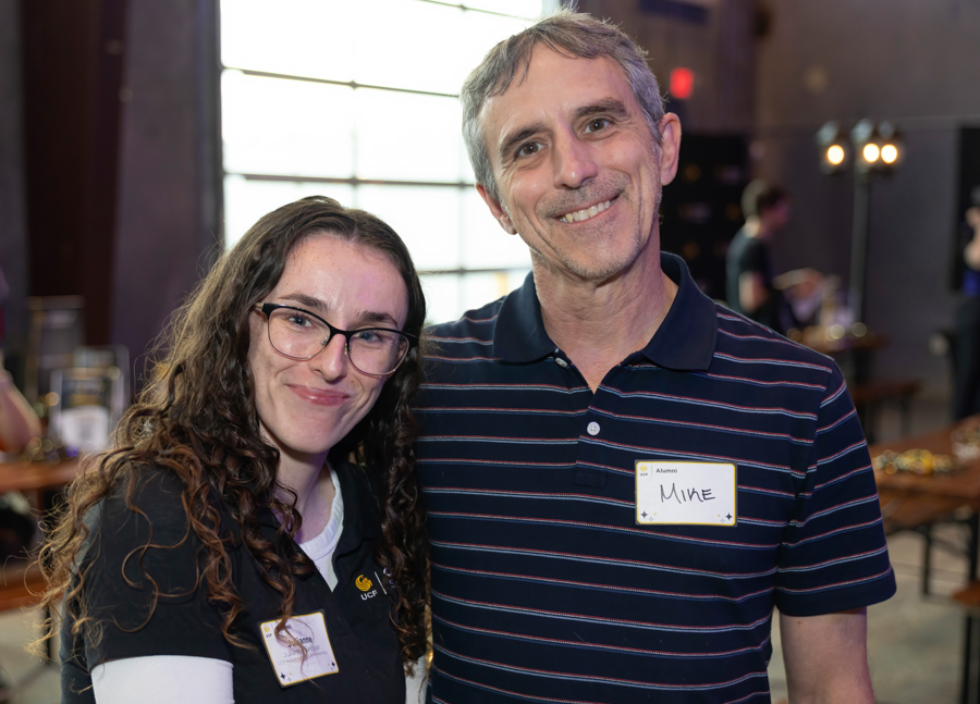 Two adults posing at a social event; woman with glasses on the left, man in a striped polo on the right, both wearing name badges.