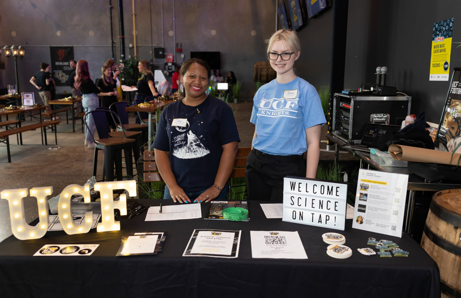 Two smiling women behind a registration table at a science event, with lit UCF letters and a 'Welcome Science on Tap' sign in view.