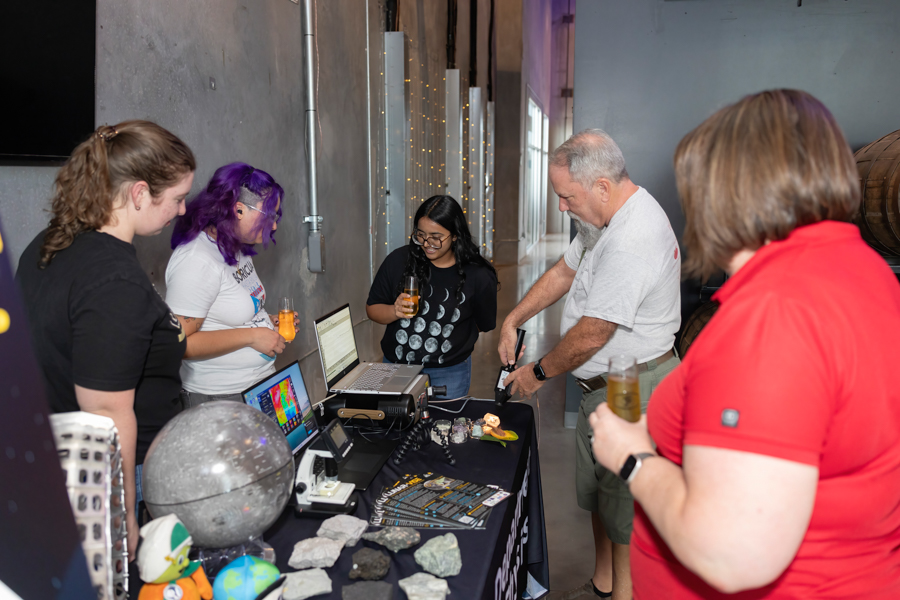 Five people stand around a demonstration table at a booth, with laptops, rocks, and brochures on display in an indoor venue.