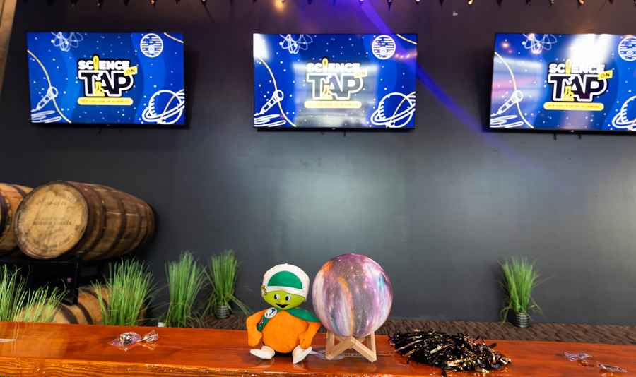 Stage area for a science event with three screens reading 'Science on Tap' against a dark wall, barrels on the left, and potted plants along the back wall.