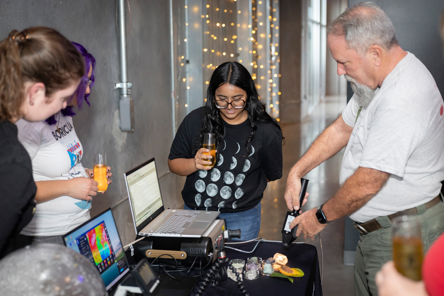 A man uses a handheld tool at a demonstration table while three people watch, each holding a glass of orange beverage and looking at laptops and small glass jars on the table.