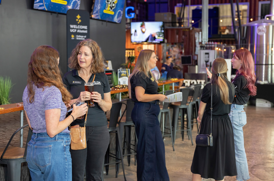 Group of women conversing at a bar in a bright event space, some holding drinks as screens and signage loom in the background.