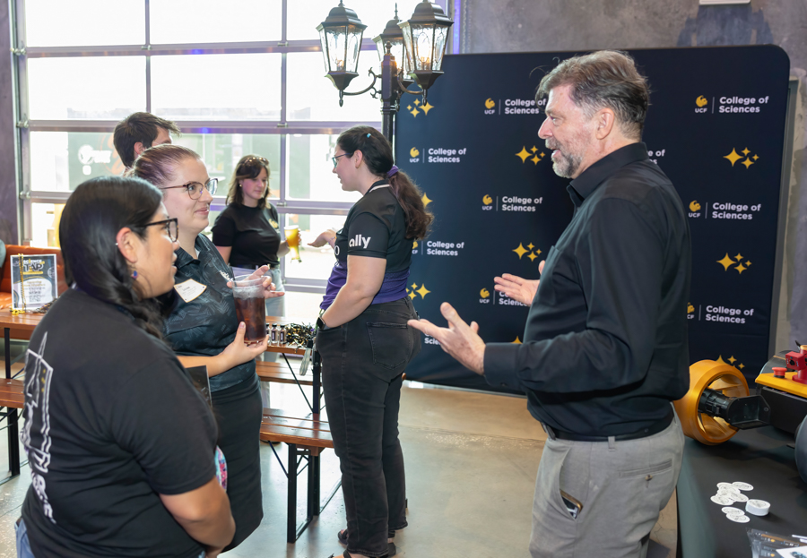 A man explains something to a small group of people at a science event, with a College of Sciences backdrop in a workshop space.