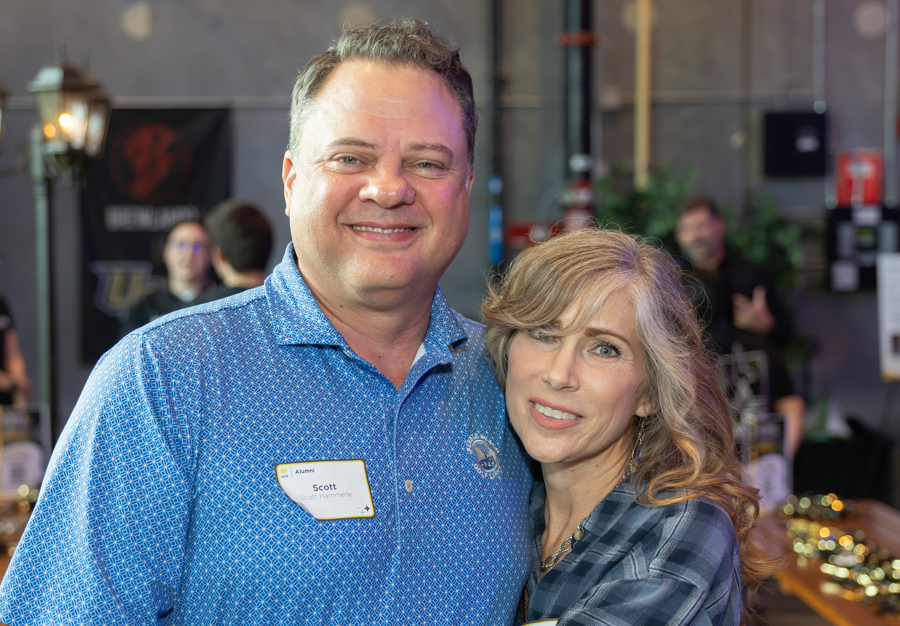 Smiling man and woman pose together at a social event, the man wearing a blue patterned shirt and a name tag while the woman leans in close behind him in a casual setting.