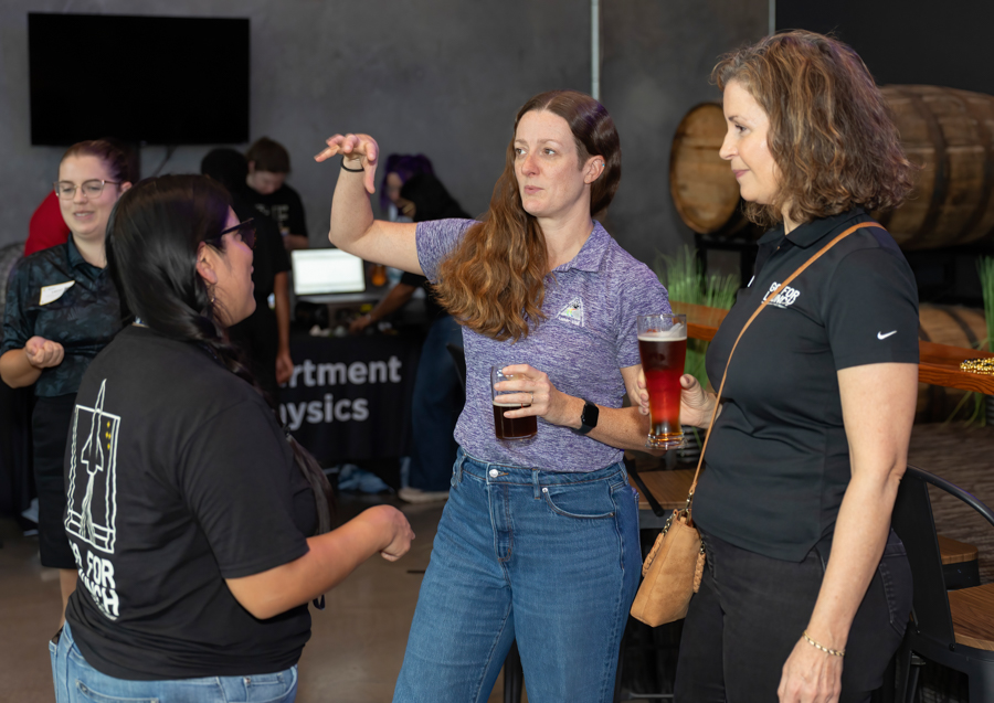Three women chat at a social event, each holding a beer, with a bar and wooden barrel in the background.