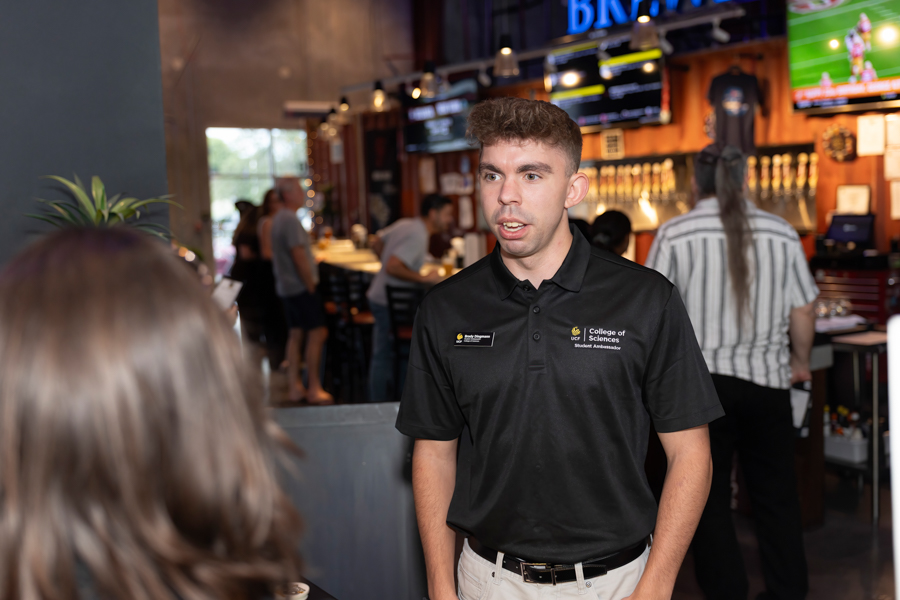 Young man in a black polo with a College of Sciences badge speaks to someone at a busy campus bar or cafe event.