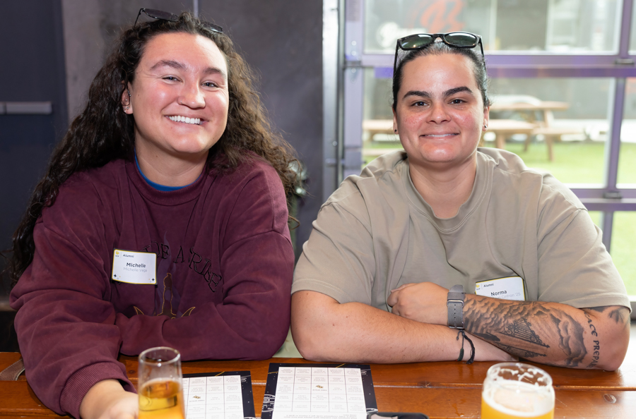 Two smiling women with name badges sit at a wooden table indoors, with drinks and menus in front.