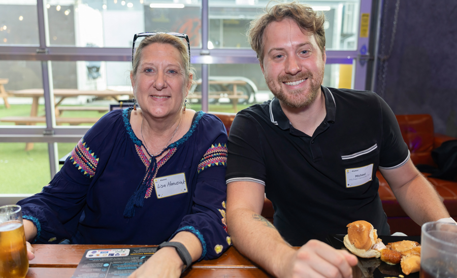 Two smiling adults sit at a wooden table indoors, wearing name tags: Lisa on the left in a blue embroidered top and sunglasses on her head, Michael on the right in a black polo; drinks and bread rolls are in front of them.