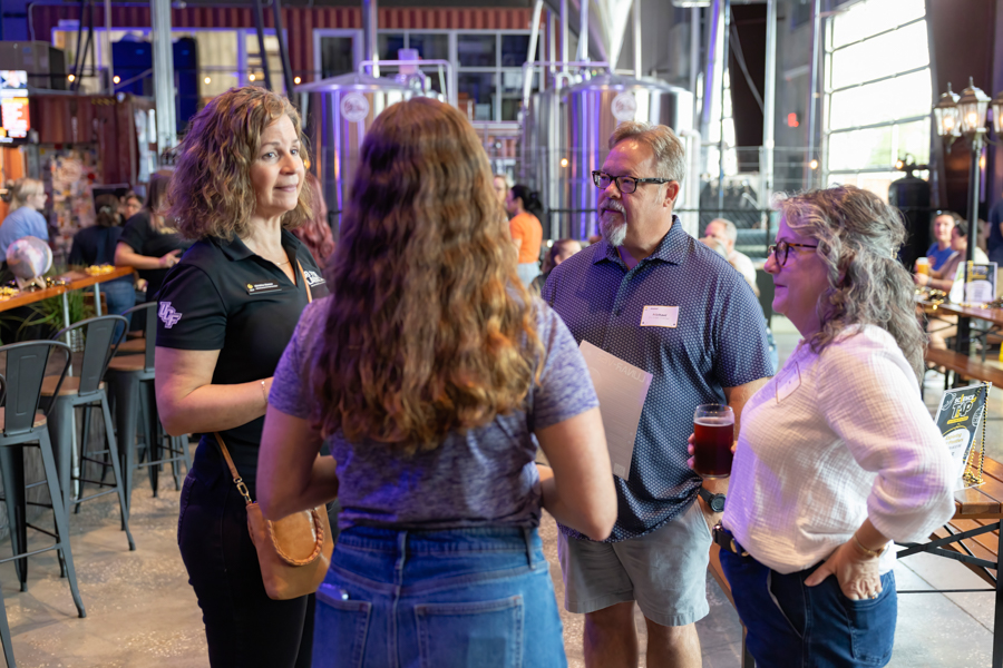 Four adults in casual attire chat at an indoor social event, with drinks in hand and brewing equipment in the background.
