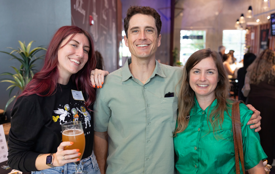 Three smiling adults posing together at a social event; a woman on the left holds a beer glass while the man in the center and a woman on the right smile.