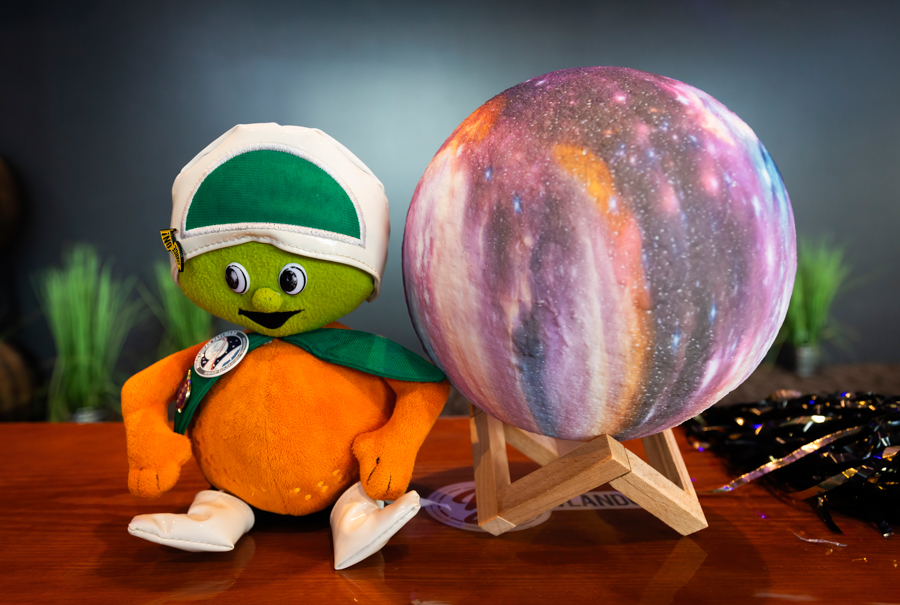 Orange plush mascot wearing a white and green helmet and green scarf sits next to a colorful planet model on a wooden stand on a table.