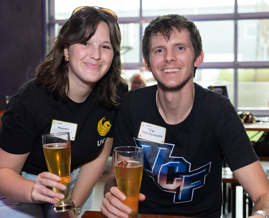 Two friends smiling at a bar, each holding a pint, with name tags visible on their shirts.