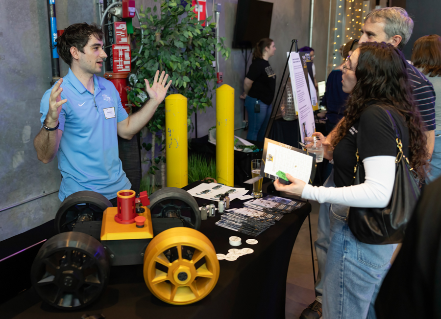 Presenter in a light blue polo explains a yellow wheel machine to visitors at a booth at a science fair/ expo.