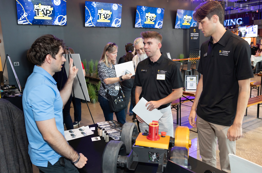 Two men at a tech expo booth discuss a machine on the table while a presenter in a blue polo explains it to them. A third person watches nearby as screens in the background display graphics.