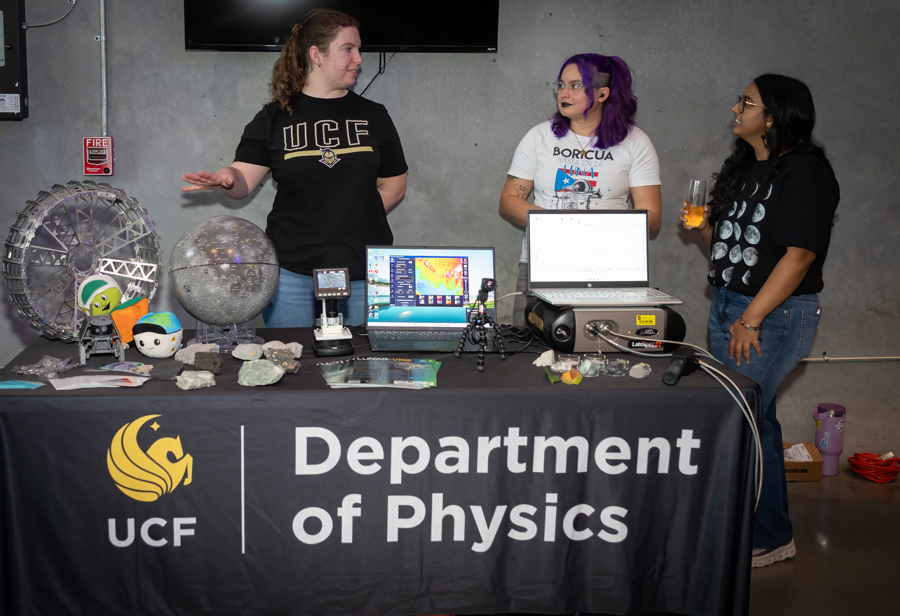 Three people stand behind a UCF Department of Physics table displaying science demos, models, and laptops, engaging in conversation.