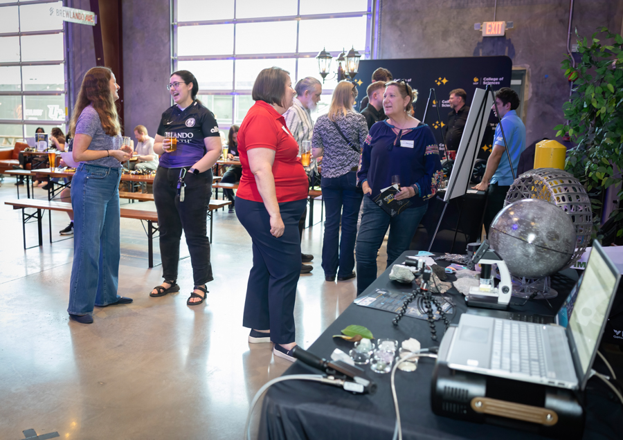 People mingle at a campus expo, chatting near exhibit tables with laptops and demo gadgets in a bright, industrial-style hall.