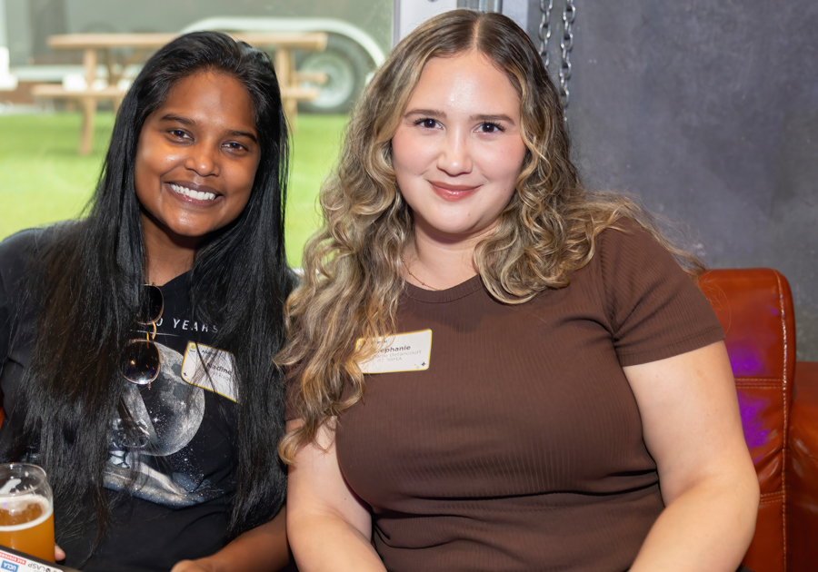 Two smiling women seated closely together for a casual photo; left has long black hair with sunglasses, right has wavy blonde hair and a name badge on a brown top.