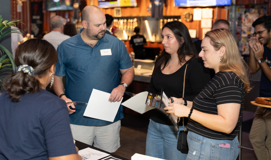 Man in a blue polo hands out brochures to two women at a registration table in a busy event space. Other attendees mingle in the background.
