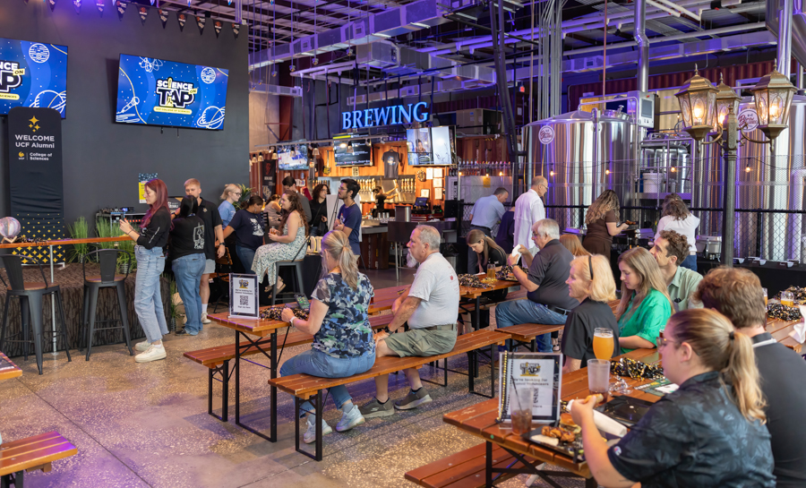 Crowd seated at long wooden benches in a modern brewery tasting room, with brewing equipment and a glowing 'BREWING' sign in the background.