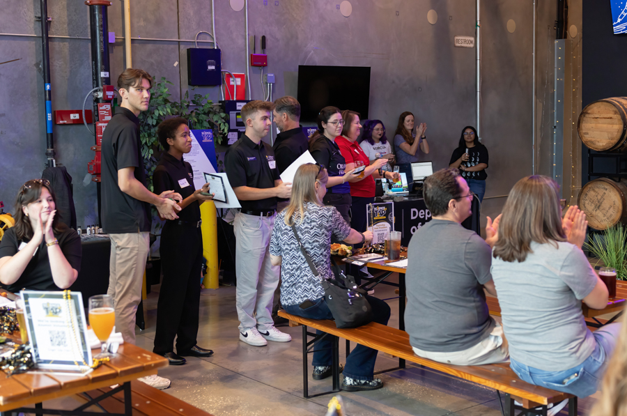 Group of people lined up to receive awards at a casual event in a brewery, with seated attendees clapping.