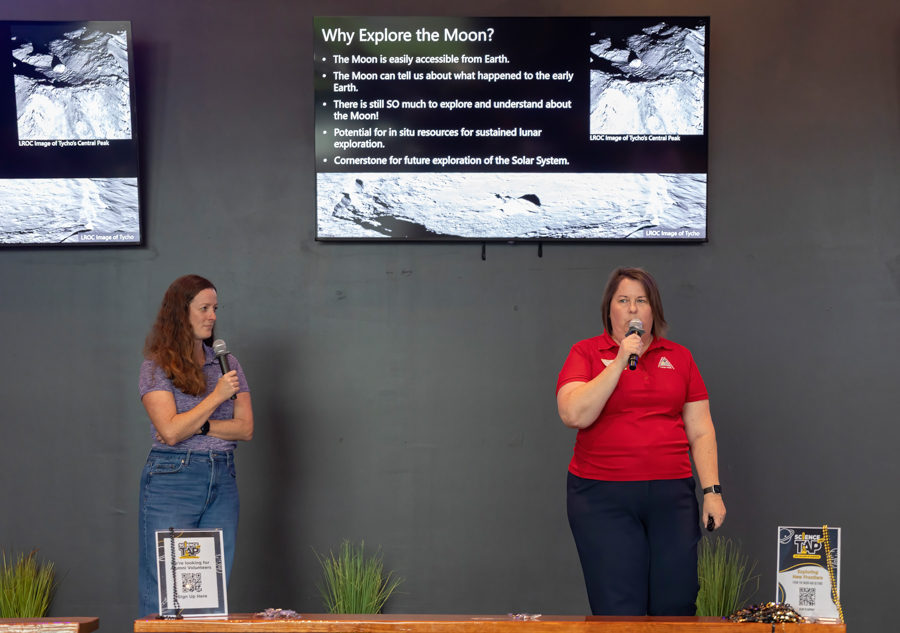 Two presenters stand on a stage giving a talk about lunar exploration; a slide reading 'Why Explore the Moon?' is projected behind them.