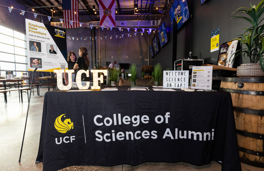 Registration table at an alumni event for the College of Sciences, with a UCF logo cloth, Welcome Science on Tap sign, posters, and two people in the background under string lights.