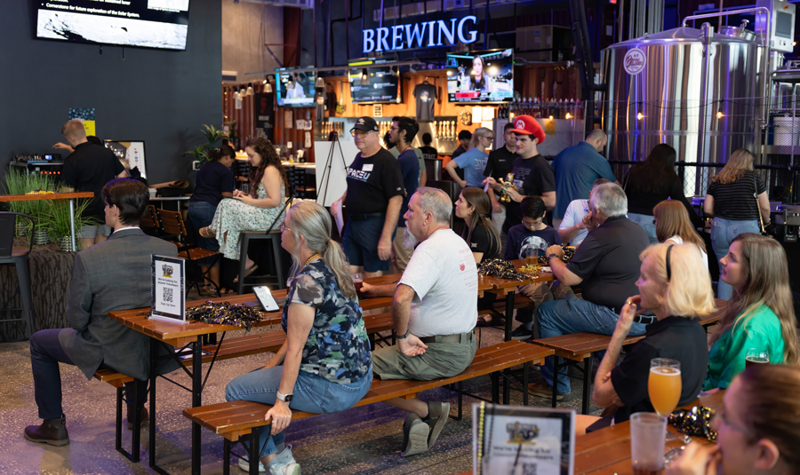 Ground-level view of a brewery tasting event: attendees sit at wooden benches, chatting and watching a presentation, with large stainless tanks in the background.