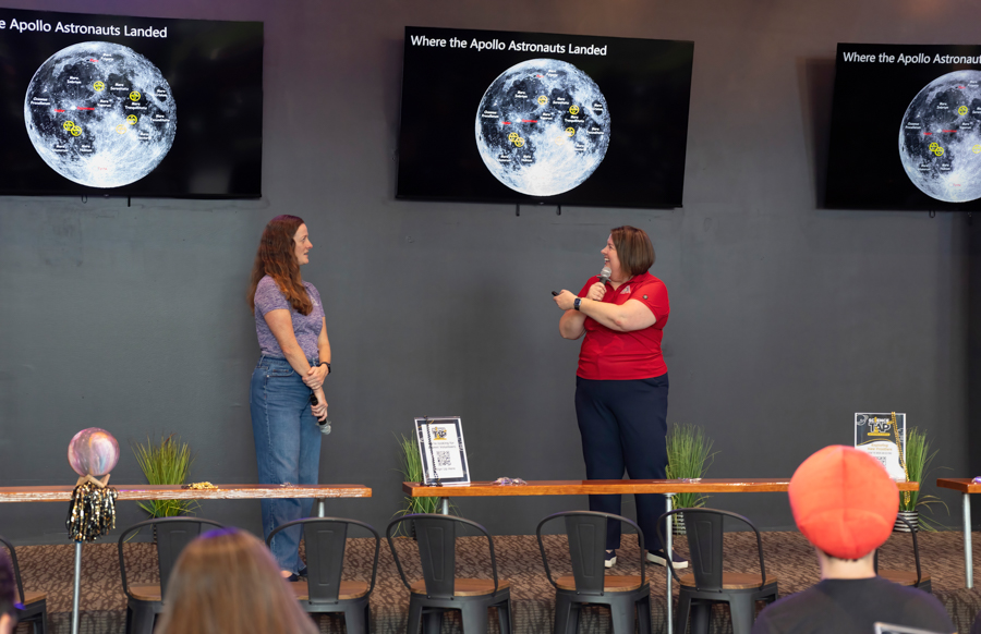 Two women on a stage giving a talk; three screens show the Moon with labeled landing sites as an audience watches.