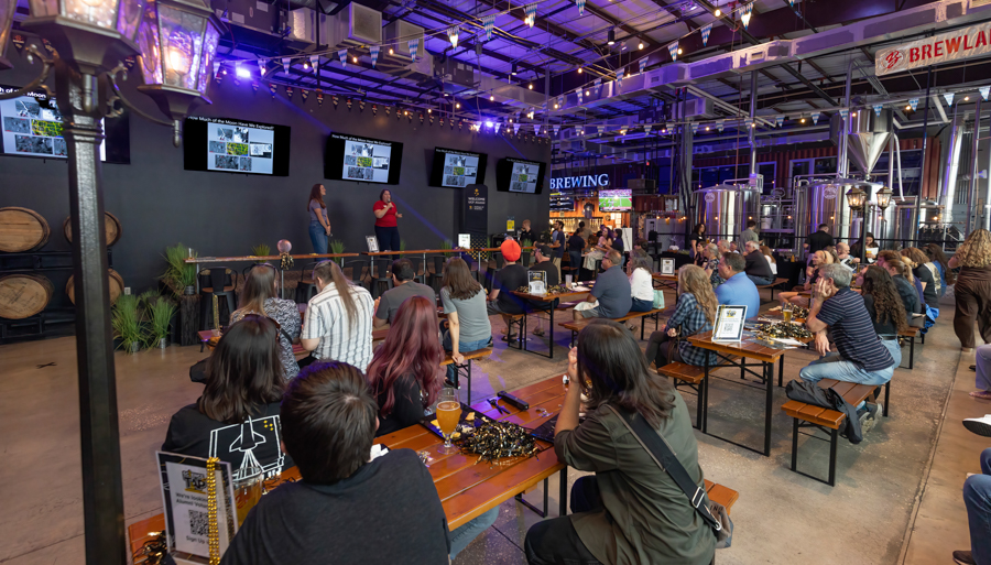 Presenter speaks on a stage in a brewery tasting room while a crowd sits at long wooden benches watching screens on the wall behind.