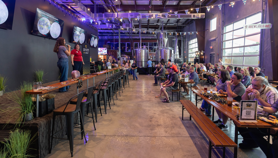 Industrial brewery hall with a stage on the left where two speakers present, row of stools along a high table, screens on the wall, and an audience at tables and benches getting beer and snacks.