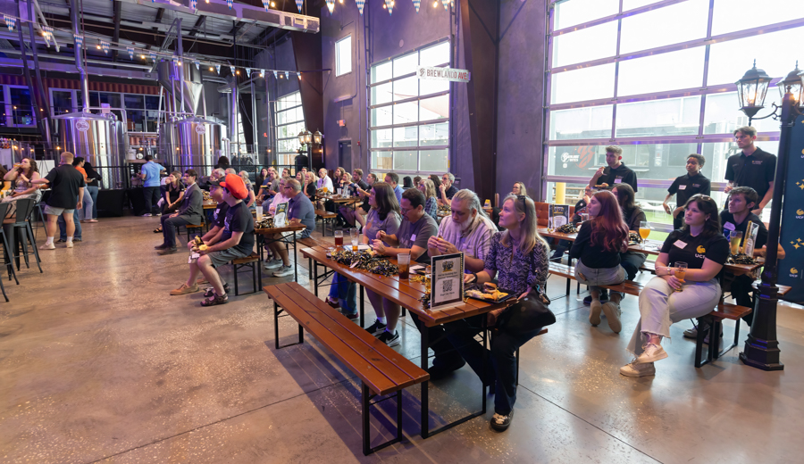 Crowd of people seated at long wooden tables inside a brewery tasting room, watching an event with brewing tanks visible in the background.