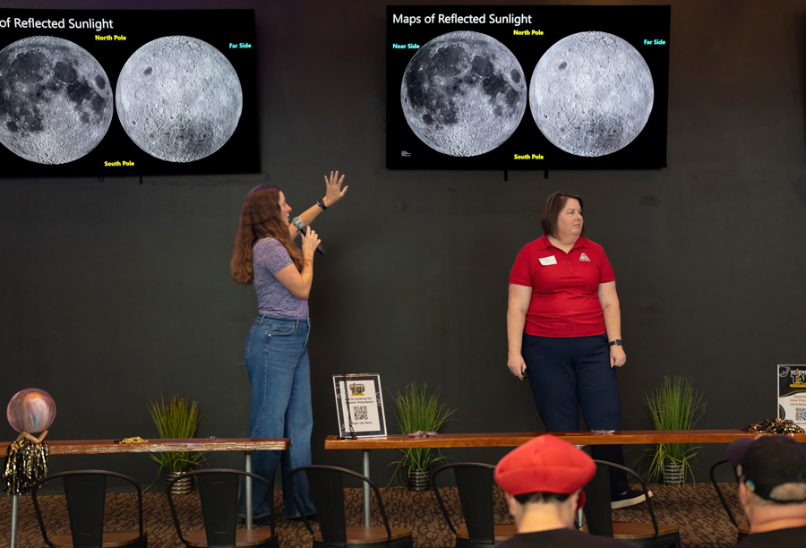 Two female presenters on a stage discussing maps of reflected sunlight, with two moon images on screens above them.