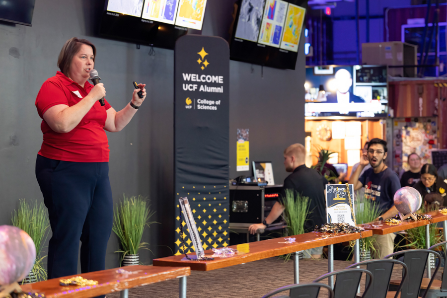 Woman in red shirt speaks into a microphone on a stage at a UCF Alumni event, with a 'Welcome' banner behind her and screens overhead.