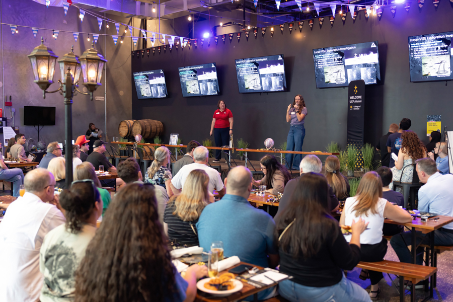 Two speakers stand on a stage in a large hall, presenting to a seated audience at long wooden tables with drinks and plates; multiple screens show the presentation behind them.