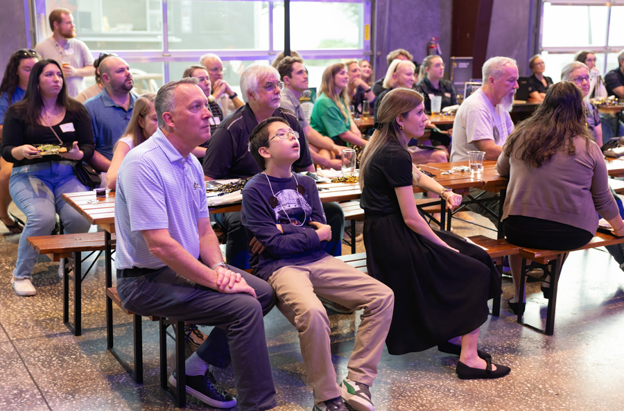Audience members seated on picnic-style benches at long tables in a bright indoor venue, watching a presentation off-camera.