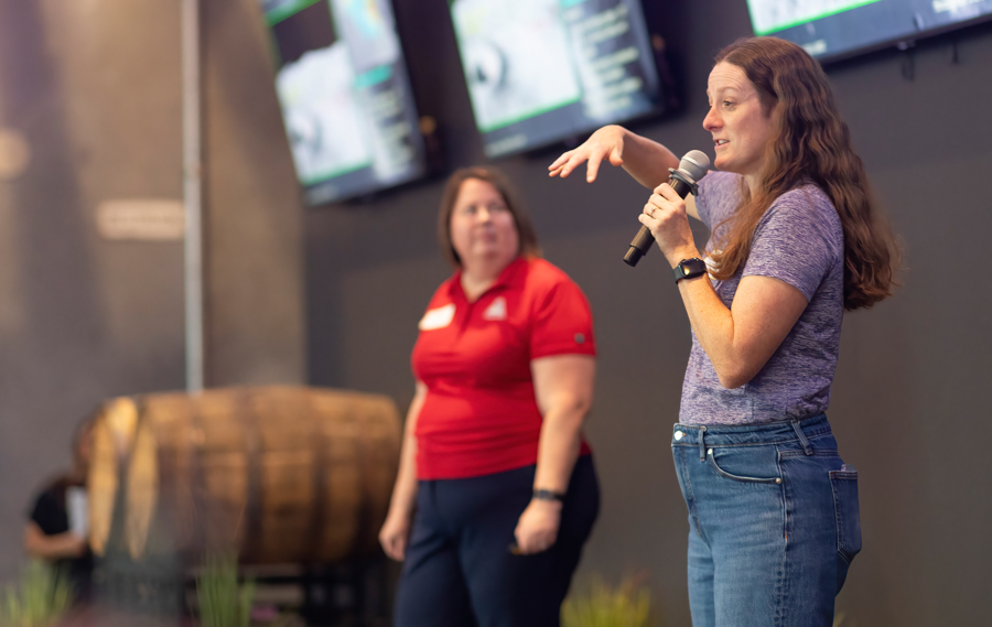 Two women on a stage: one speaks into a microphone while gesturing, the other in a red polo watches, with screens in the background.