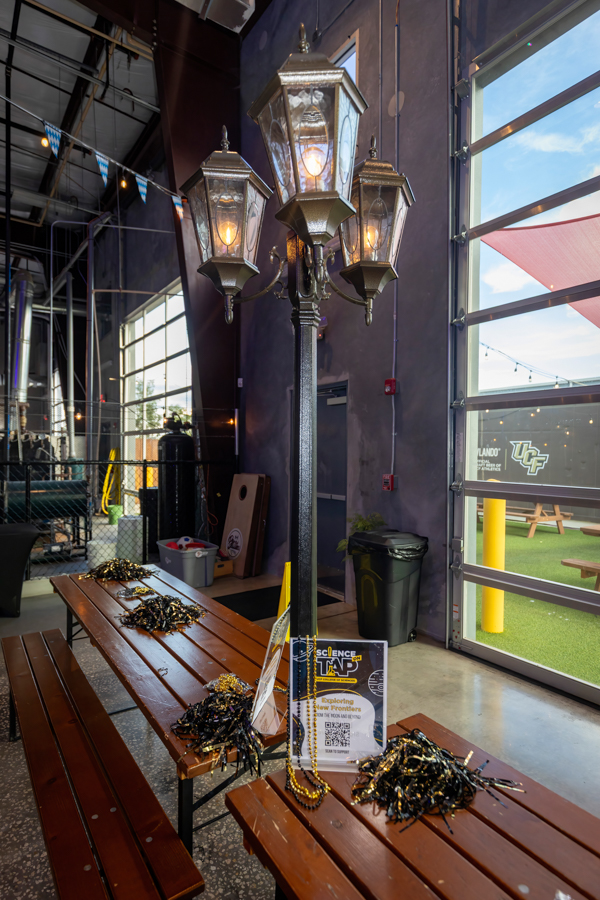 Indoor science-event space featuring a tall decorative street lamp, wooden benches, and black-and-gold bead garlands on the tables beside a large open garage door.