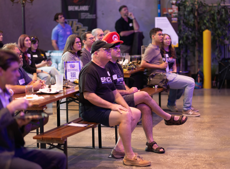 Audience seated at a long wooden table during an indoor event, one person wearing a red Mario cap watching the stage. Hint: event or meetup setting with attendees listening to a presentation.
