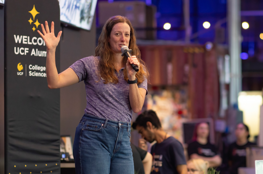 Woman on stage speaks into a handheld mic, waving to the audience at a UCF Alumni event with a Welcome sign in the background.